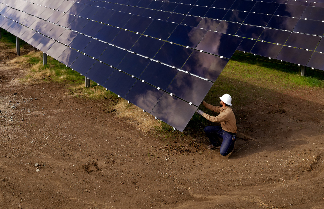 Funcionário com capacete de segurança inspeciona painéis solares em um campo.